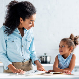 cheerful african american mother and daughter looking at each other, cookbook on table in kitchen