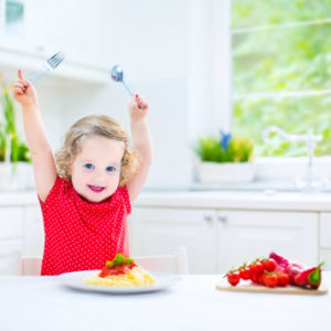 Cute curly laughing toddler girl in a red shirt playing with fork and spoon eating spaghetti with tomato sauce and vegetables for healthy lunch sitting in a white sunny modern kitchen with big window with ideas for picky eaters - SuperKidsNutrition.com