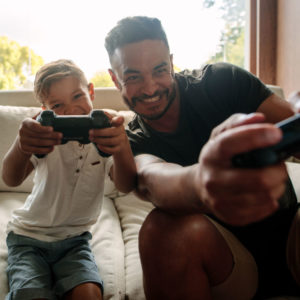 Portrait of father and son enjoying playing video game in living room. Smiling young man and little boy playing video game at home.