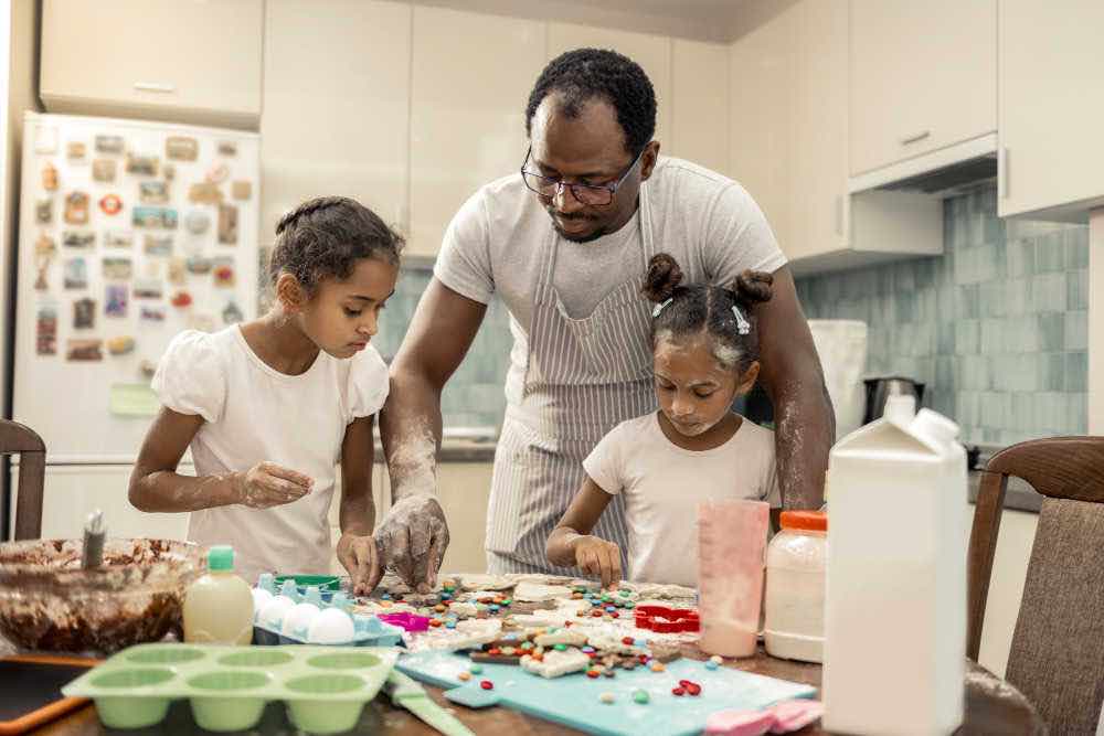 Helping girls. Caring loving father in striped apron helping his girls making cookies for gifts