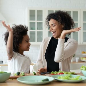 Excited African mom give high five gesture to little adorable mixed-race daughter family cook together dinner healthy food vegetarian salad. Teach kid, happy motherhood, share cookery skills concept to prevent colon cancer starting young